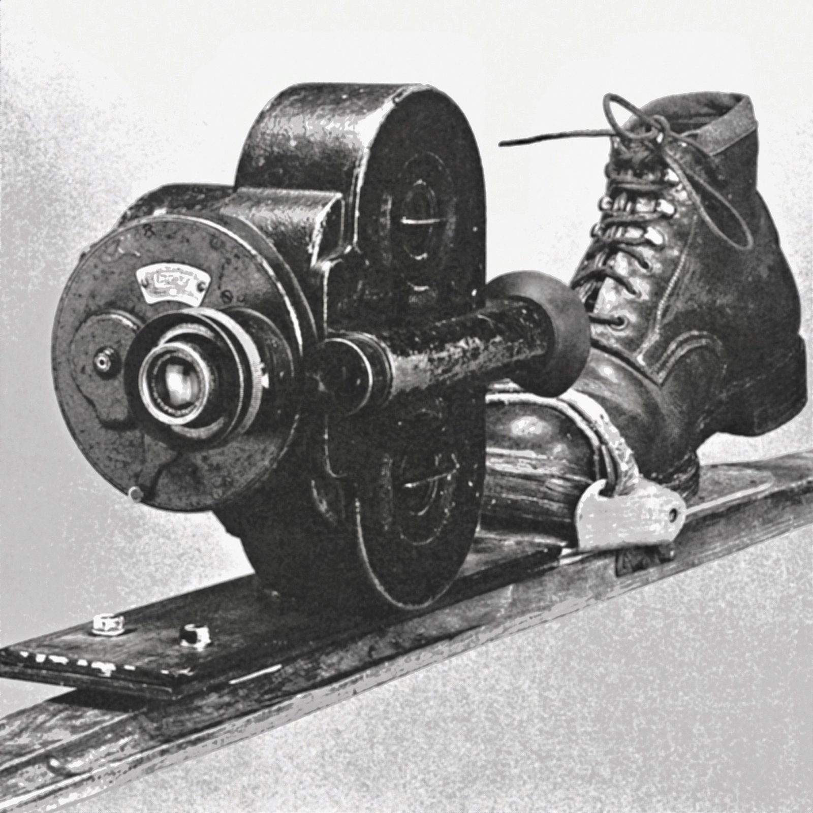 A Bell & Howell Eyemo camera bolted to a ski alongside a laced boot in a binding, photographed in black and white.