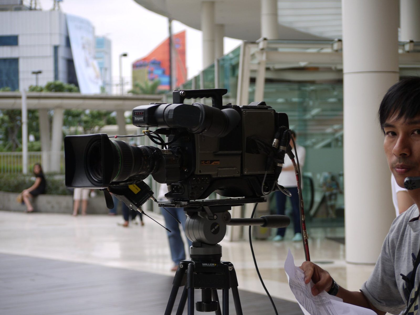 An Ikegami HL-45 broadcast camera with a zoom lens on a tripod outside a building, with a crew member in the foreground.