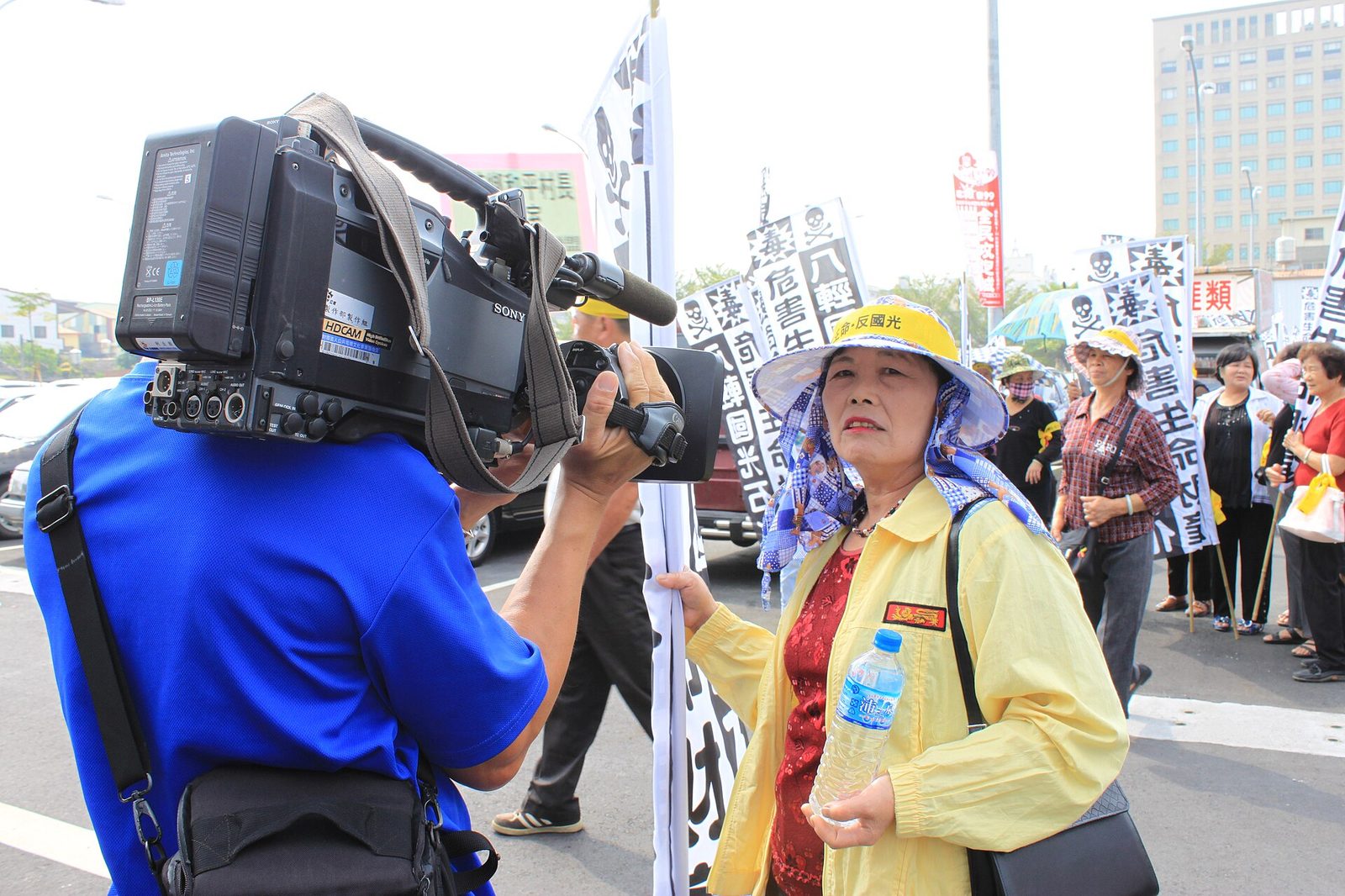 A news cameraman shoulders a Sony HDCAM camcorder while filming a woman at a street protest.