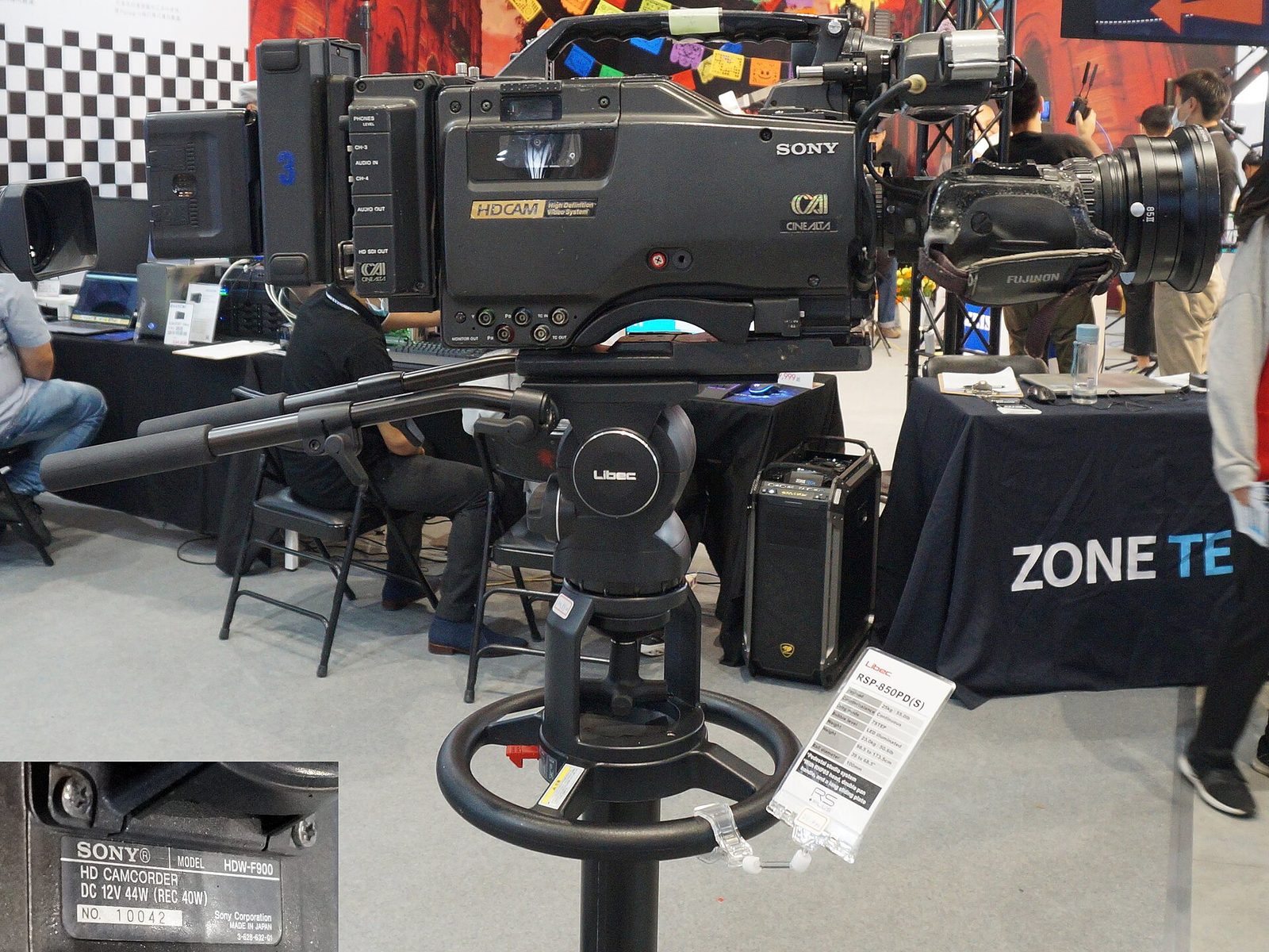 A Sony HDW-F900 CineAlta camcorder on a Libec tripod head at a trade show booth, with its model plate shown in an inset.