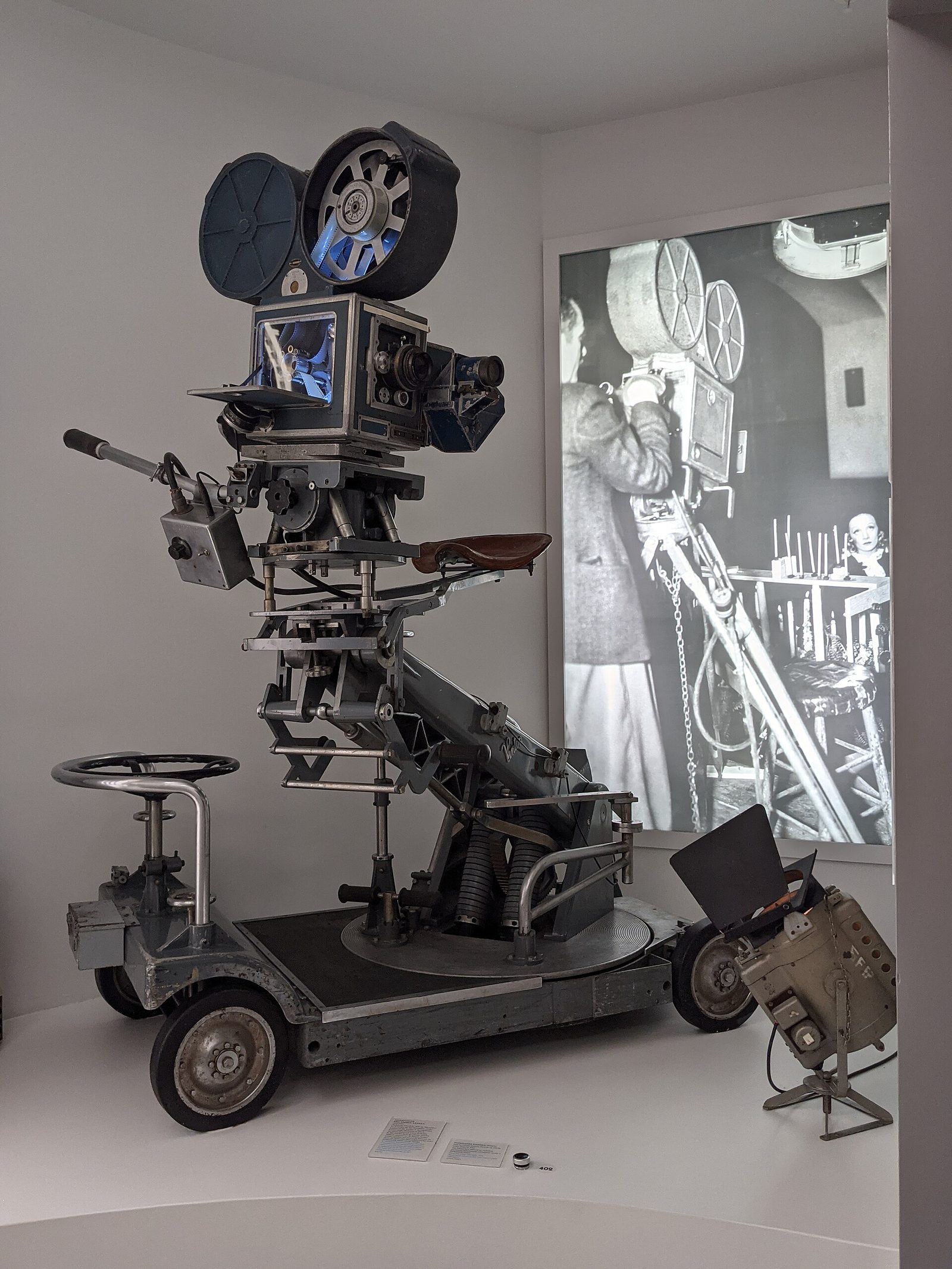 A Technicolor three-color camera mounted on a wheeled Pathfinder dolly in a museum gallery, with a large period photograph of a film crew on the wall behind it.