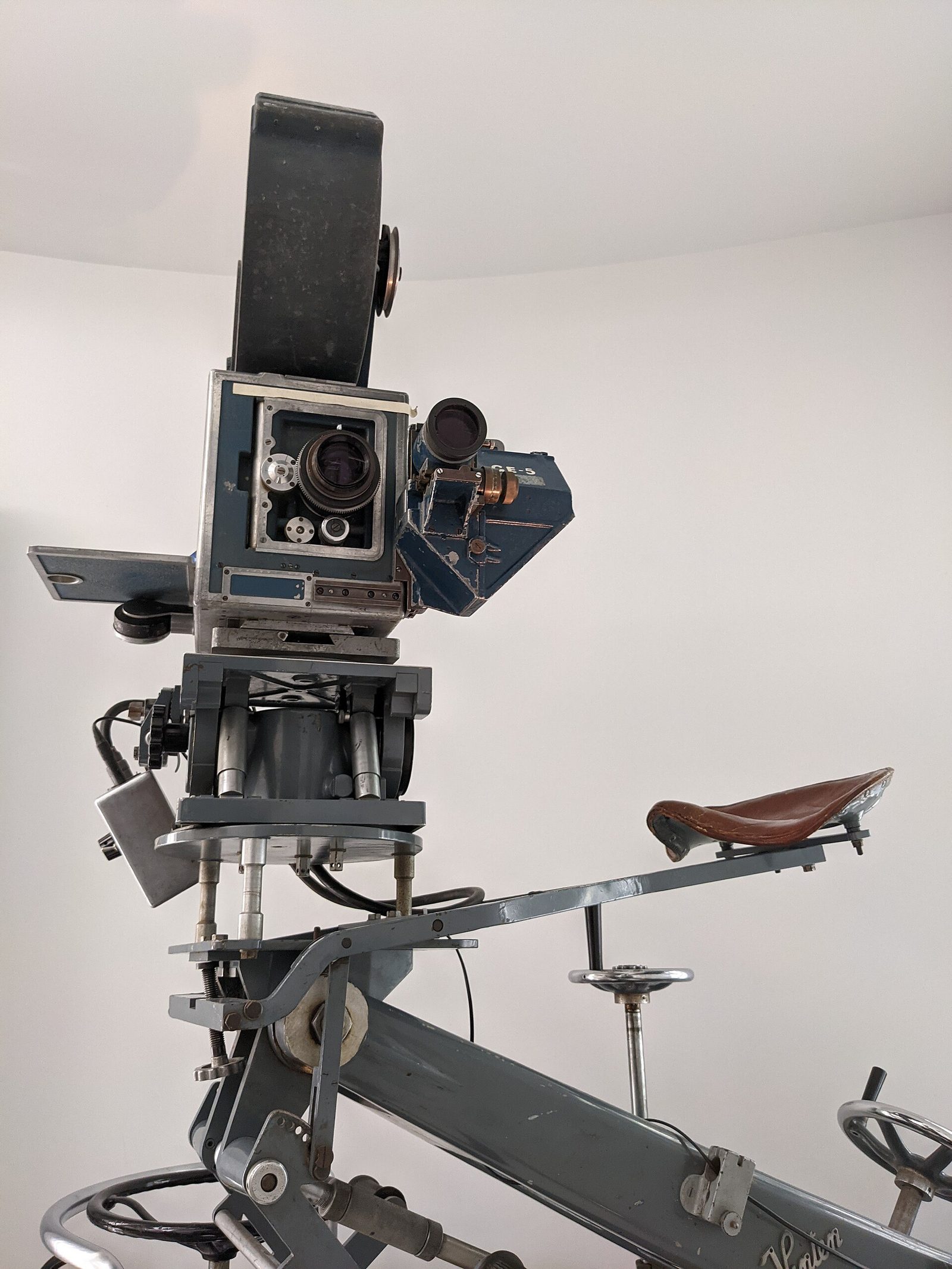 Close view of the same Technicolor camera with its front panel open, showing the lens port, the separate optical viewfinder beside it, and a leather bicycle seat on the dolly arm.